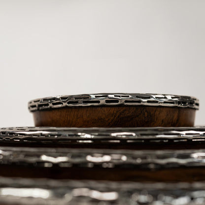 Nahaufnahme der oberen Ränder von ineinander verschachtelten Schalen aus Teakholz, die mit einem dekorativen silbernen Metallgitter umrandet sind. Die Kamera fokussiert sich auf das metallische Muster, das im scharfen Kontrast zum warmen Braun des Holzes steht.